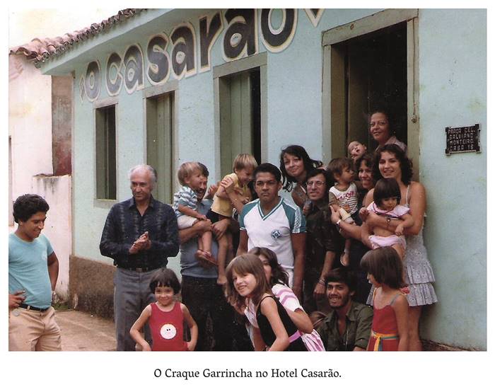  4 e. Foto colorida de frente ao hotel Casarão, onde o jogador de Futebol Mané Garrincha posa ao lado de homens, mulheres e crianças que sorriem. Mané Garrincha é um homem negro, cabelos curtos, ele veste camiseta verde e branca e short branco. Realização: Academia Planaltinense de Letras, Artes e Ciências (APLAC), pelo Ecomuseu Pedra Fundamental e pelo Coletivo Nativo Audiodescrição produzida  pelo Instituto de Promoção das Pessoas com Deficiência Visual Audiodescritora: Elma Lúcia Rodrigues Consultor: Fernando Rodrigues Este projeto é promovido com recursos do Fundo de Apoio a Cultura do DF